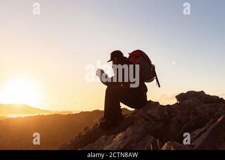 Side view of unrecognizable male hiker sitting on rock in highlands and taking photo while admiring sunset Stock Photo