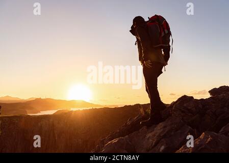 Side view of unrecognizable male traveler with backpack standing on hill and admiring scenery of mountain ridge under sunset sky Stock Photo