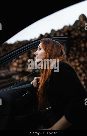 Low angle side view of young Woman standing near open door of car against blurred stacked wooden logs and looking away while travelling through countryside in autumn day Stock Photo