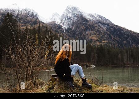 Side view of unrecognizable distant female traveler in warm outfit sitting on rocky shore of lake and observing amazing scenery of autumn forest and snowy mountains in cloudy day Stock Photo