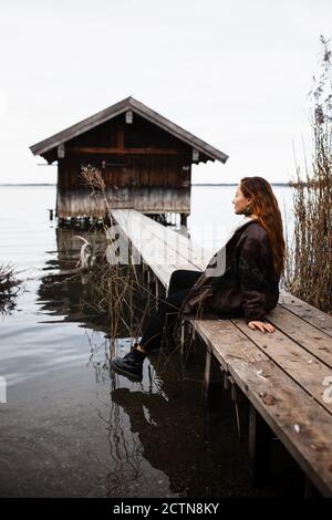 Female traveler in warm clothes sitting on wooden plank pier near shabby shed located at lake in gloomy autumn day Stock Photo