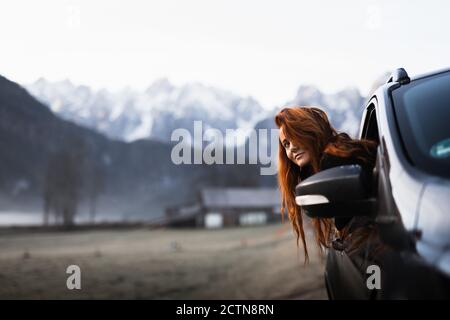 Side view of young female tourist looking out from open window of car while travelling through autumn countryside with rocky mountains covered by snow Stock Photo