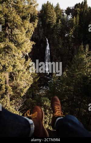Crop unrecognizable traveler in boots sitting on mountain slope against waterfall streaming among dense green coniferous forest in USA Stock Photo