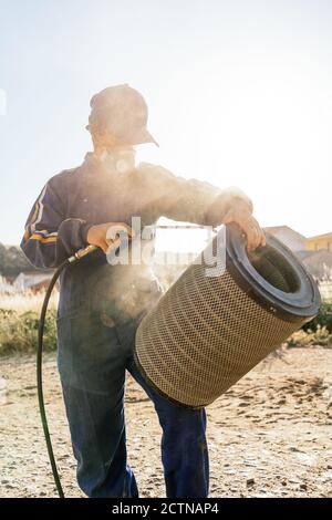 Unrecognizable farmer in workwear and protective respirator mask spraying water from hose to dirty filter of agricultural machine during maintenance work in farm yard Stock Photo