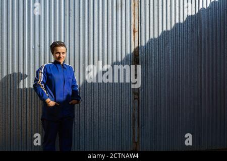 Cheerful female worker in uniform standing near metal wall of building in rural area and looking at camera Stock Photo
