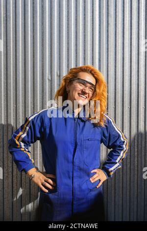 Cheerful female engineer in protective glasses and workwear standing near metal wall at plant and looking away while enjoying break during work Stock Photo