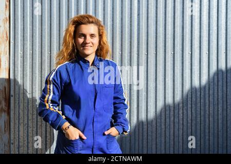 Cheerful female engineer in protective glasses and workwear standing near metal wall at plant and looking at camera while enjoying break during work Stock Photo