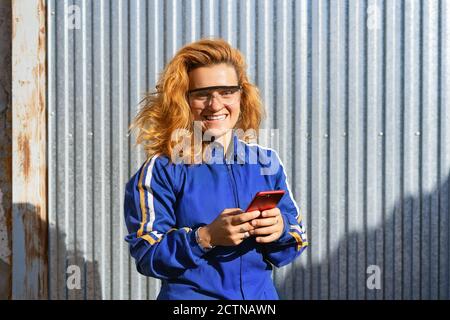 Delighted female worker in uniform and protective glasses standing near metal building and browsing cellphone while having break during work Stock Photo