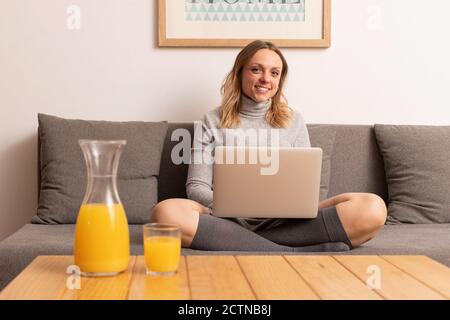 Smiling female freelancer sitting on sofa in living room and working on remote project while using netbook and looking at camera Stock Photo