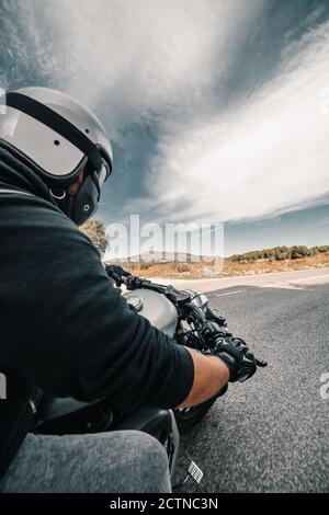 Back view cropped unrecognizable male rider in leather jacket and helmet riding motorbike on roadside against cloudy sky in countryside Stock Photo