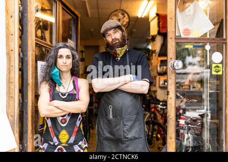 Confident adult ethnic man and Woman in workwear with protective masks standing on doorway of bicycle service workshop and looking at camera Stock Photo