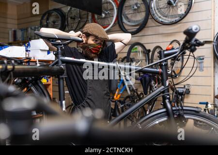 Serious bearded male master in workwear putting on protective mask while working in bicycle service workshop during coronavirus pandemic Stock Photo