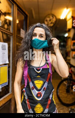 Positive adult ethnic female mechanic in colorful apron with protective mask smiling and looking at camera while standing on entrance of bike service workshop Stock Photo