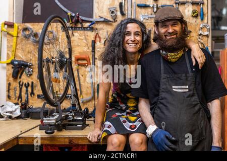 Positive ethnic couple of owners of bicycle service workshop looking at camera and smiling friendly against weathered wall with various repair instruments Stock Photo