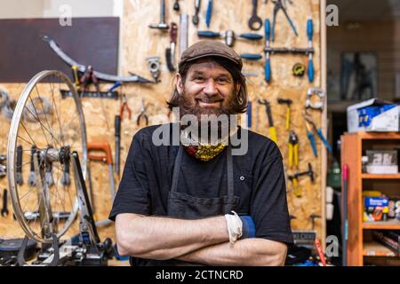 Smiling middle aged bearded craftsman in workwear looking at camera friendly while standing against weathered workbench with various tools in bicycle service workshop Stock Photo
