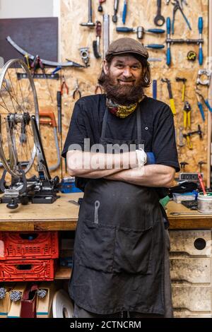 Smiling middle aged bearded craftsman in workwear looking at camera friendly while standing against weathered workbench with various tools in bicycle service workshop Stock Photo