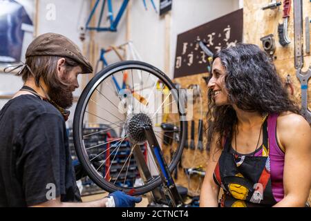 Side view of focused bearded man fixing bike wheel and cheerful Woman sitting nearby while working together in workshop Stock Photo