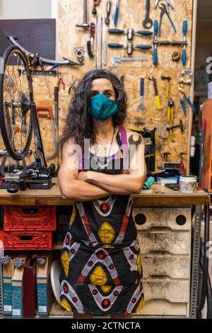 Serious adult ethnic female mechanic in workwear and protective mask holding wrench and looking at camera while standing against weathered workbench with various tools in bicycle service workshop Stock Photo