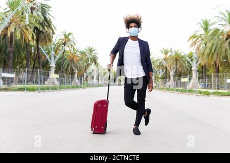 Young black man in a suit standing looking at camera with wheeled suitcase on the street on a tree-lined avenue Stock Photo
