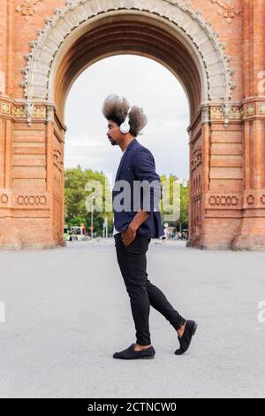 Side view of black man with afro hair and a casual suit walking down the street while listening to music with headphones Stock Photo