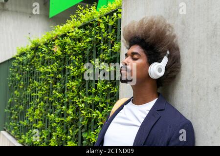 Black man with afro hair leaning against a wall with his eyes closed while listening to music with a wireless headset on the street Stock Photo