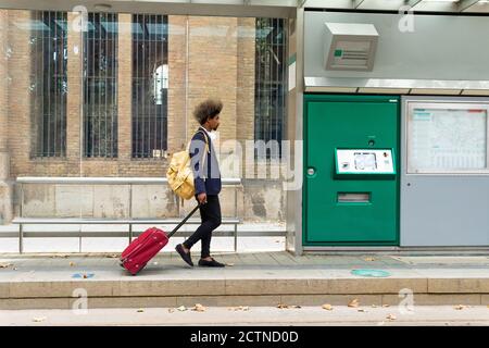 Side view of black man with afro hair walking on a tram station with a suitcase on wheels and a bag hanging from his shoulder Stock Photo