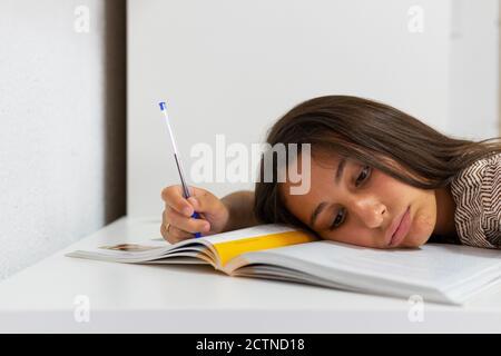Exhausted female student lying on table with textbook and pen and sleeping while preparing for exam Stock Photo