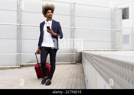 Black man in a suit with a wireless headphones around his neck and a suitcase walking while using the mobile outdoors Stock Photo
