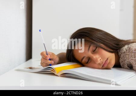 Exhausted female student lying on table with textbook and pen and sleeping while preparing for exam Stock Photo