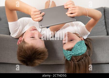 Brother and sister lying face down on the couch while entertained using tablet during weekend Stock Photo