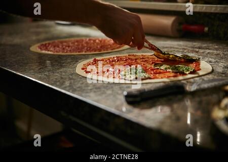 Top view of anonymous chef pouring and spreading tomato sauce on dough with ladle while cooking pizza on table in restaurant Stock Photo