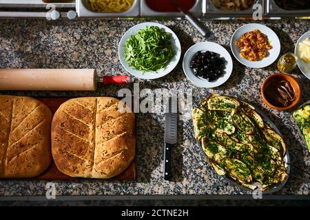Top view of baking tray with hot freshly baked focaccia bread on marble table with various ingredients in restaurant kitchen Stock Photo