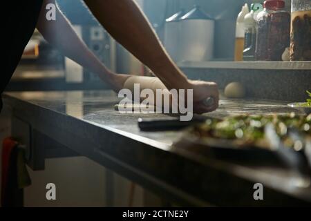 Cropped anonymous man using rolling pin to stretch fresh dough on on marble counter table while preparing pizza in kitchen Stock Photo