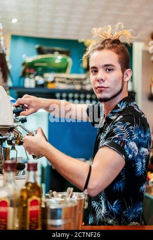 Side view of cheerful male barista with dreadlocks preparing aromatic coffee in coffee maker while looking at camera Stock Photo