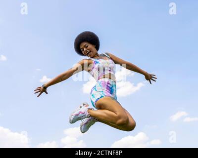 Low angle of delighted African American female in summer outfit in moment of jumping above ground on background of blue sky