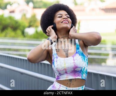 Carefree African American female listening to music in wireless headphones and enjoying songs with closed eyes while standing on street in summer Stock Photo