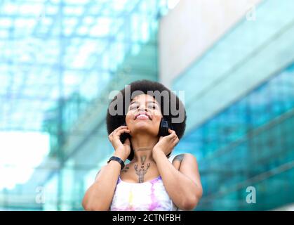 Carefree African American female listening to music in wireless headphones and enjoying songs with closed eyes while standing on street in summer Stock Photo