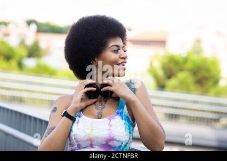 Carefree African American female listening to music in wireless headphones and enjoying songs while standing on street in summer Stock Photo