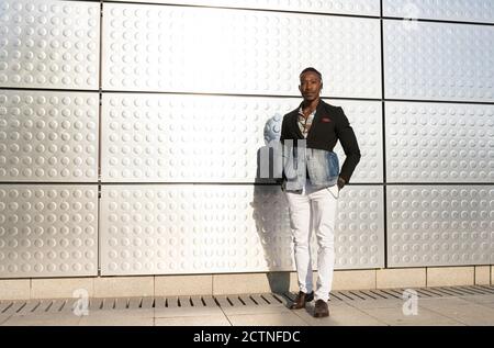 Serious African American male model in fancy outfit standing near silver wall on street and looking at camera Stock Photo