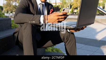 Crop african American businessman sitting on steps with laptop in city and reading messages on cellphone while working remotely Stock Photo
