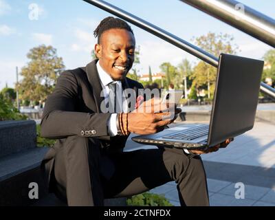 Cheerful African American businessman sitting on steps with laptop in city and reading messages on cellphone while working remotely Stock Photo