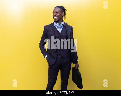 Stylish African American businessman in suit and with briefcase standing on yellow background in city and looking away Stock Photo