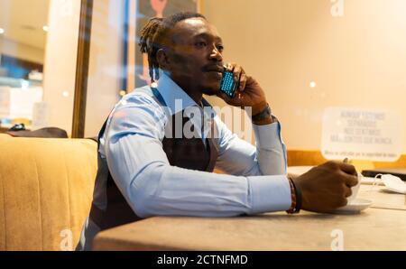 Serious African American male entrepreneur in classy outfit sitting at table with cup of coffee in cafe and speaking on cellphone while looking out of window Stock Photo