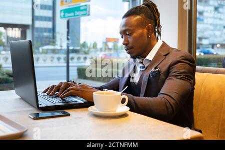 Side view of concentrated African American businessman in suit sitting at table with coffee in cafe and typing on netbook while working on remote project Stock Photo