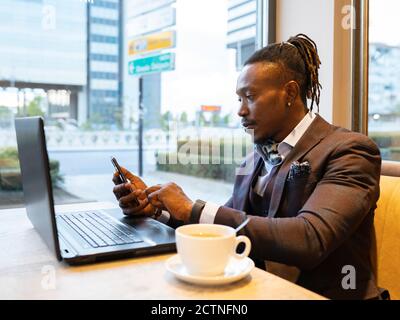 Side view of concentrated African American businessman in suit sitting at table with coffee in cafe and browsing smartphone while working on remote project Stock Photo