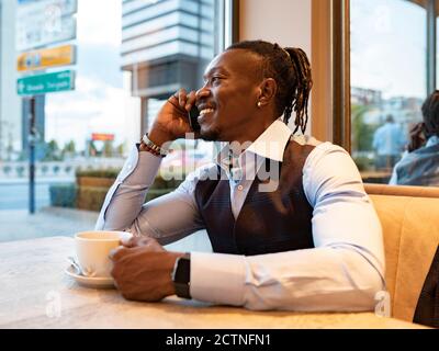 Positive African American male entrepreneur in classy outfit sitting at table with cup of coffee in cafe and speaking on cellphone while looking out of window Stock Photo