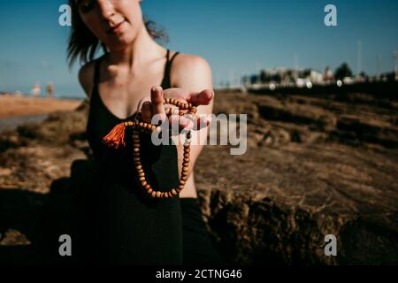 Calm crop female sitting in yoga pose on rocky shore and showing prayer beads