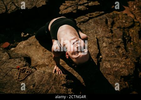 From above of calm female practicing yoga in Ustrasana with closed eyes on rocky shore in summer Stock Photo