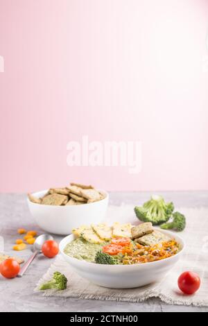 Green broccoli cream soup with crackers and cheese in white bowl on a gray and pink background and linen napkin. side view, copy space. Stock Photo