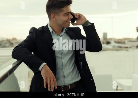 Smiling male entrepreneur in formal suit leaning on metal railing in city and talking on smartphone while looking away Stock Photo